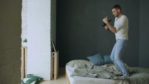 Young man singing to hair dryer and dancing hiphop on bed in bedroom - Materiał filmowy, wideo