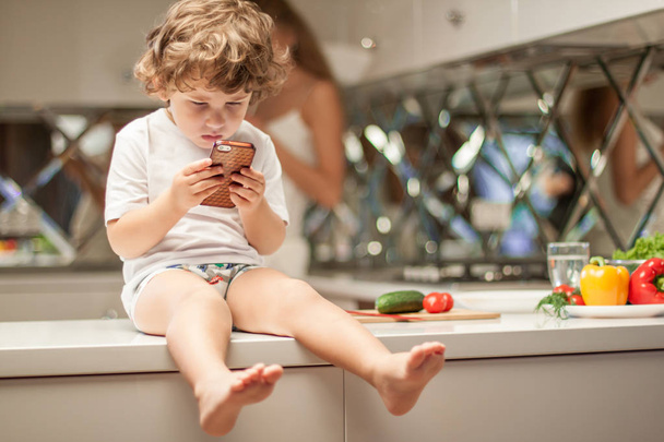 Lindo niño pequeño sentado en la superficie de la cocina con el teléfono en sus manos
. - Foto, Imagen