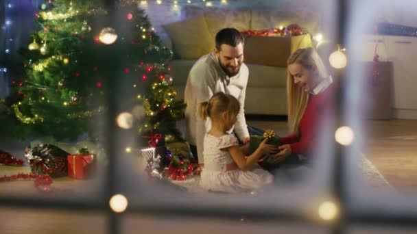 Looking Through Snowy Window. Happy Father, Mother and Daughter Sitting Under Christmas Tree. Daughter Gives a Gift to Her Mother. - Filmagem, Vídeo