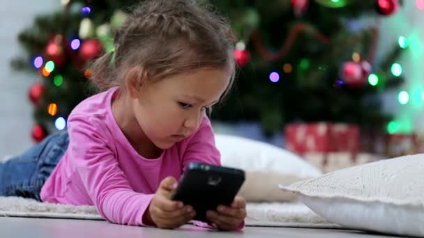 Little cute girl using smart phone, in front of Christmas tree. Close-up shot. - Video, Çekim