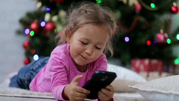 Little cute girl using smart phone, in front of Christmas tree. Close-up shot. - Filmati, video