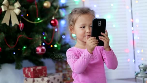 Little cute girl making selfie with Christmas tree on the background - Metraje, vídeo