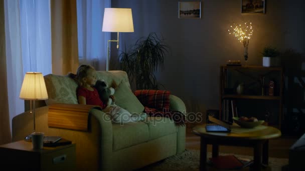 In the Evening Cute Little Girl is Sitting in Living Room on a Sofa, She Watches Television. She Holds Her Soft Toy, Room Lights are On. - Video, Çekim