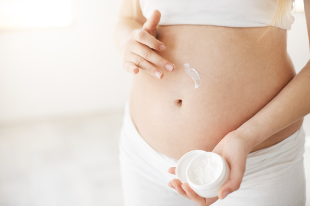 Close up of a happy pregnant lady using anti stretch mark cream to hydrate her healthy tummy. Prenatal care concept. High key. - Фото, изображение