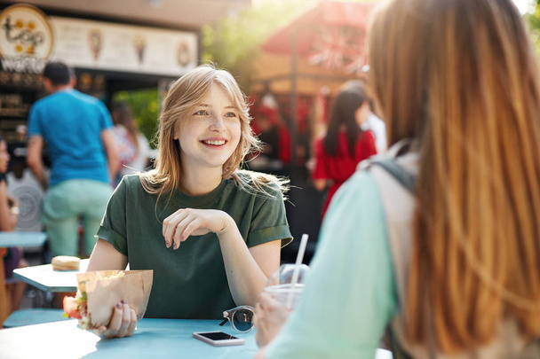 beste Freunde plaudern an einem sonnigen Tag Tacos essen im Park oder auf einem Jahrmarkt, lächeln fröhlich und diskutieren über ihr zukünftiges Start-up für gesunde Ernährung. - Foto, Bild