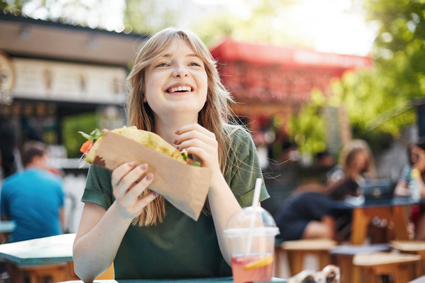 Girl eating taco. Hungry freckled blonde woman holding junk food on a food court on a sunny summer day in park smiling off camera enjoying her meal. - Photo, Image