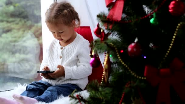 Decorated Christmass tree and little girl with smart phone on the background. - Imágenes, Vídeo