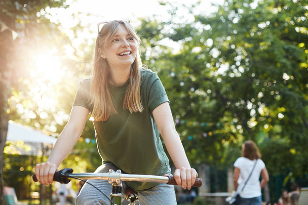 Portrait of young beautiful blonde woman enjoying pretending to ride a bicycle in the park during a food festival smiling off camera - Fotografie, Obrázek