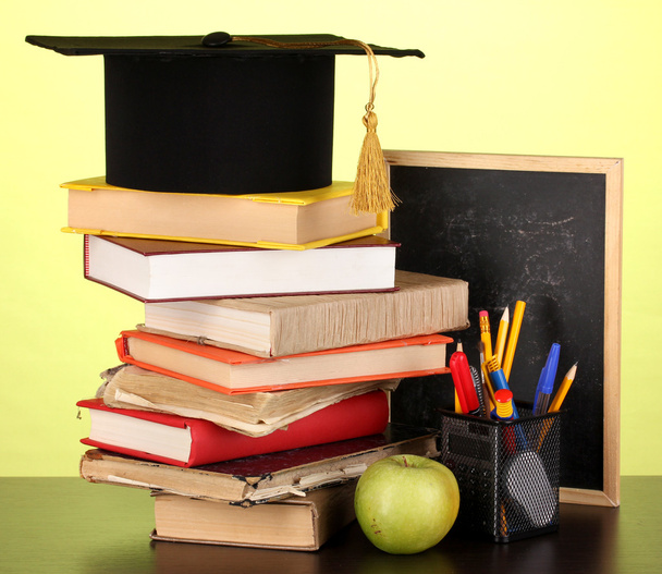 Books and magister cap against school board on wooden table on green background - Photo, Image