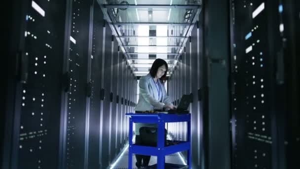 Азиатский инженер по информационным технологиям работает над портативным компьютером в большом центре обработки данных, полном стеллажных серверов
. - Кадры, видео