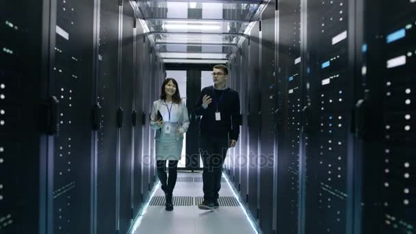 Techniciens informatiques caucasiens masculins et asiatiques marchant dans un centre de données avec des rangées de serveurs de rack. Ils ont une discussion, elle tient Tablet Computer
. - Séquence, vidéo