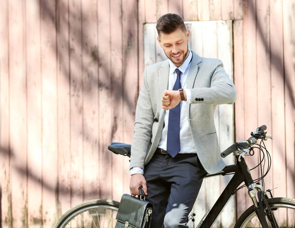 Przystojny, młody biznesmen, patrząc na zegarek, stojąc w pobliżu rower na zewnątrz w słoneczny dzień - Zdjęcie, obraz