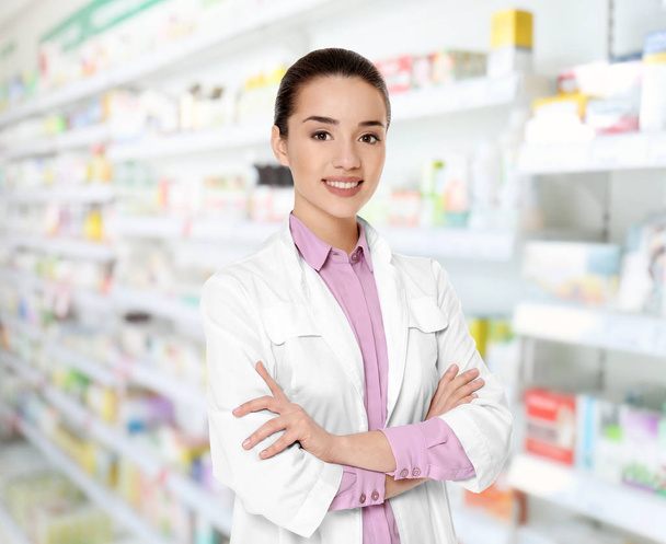 Young female pharmacist at work. Blurred shelves with pharmaceutical products on background - 写真・画像