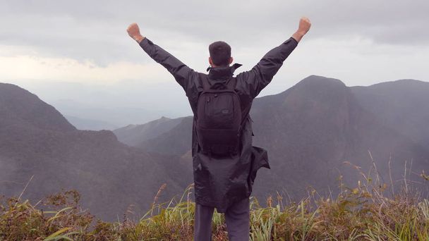 Молодой турист в плаще с рюкзаком поднимается на вершину горы и поднимает руки. Человек-турист, стоящий на краю красивого каньона, победно протягивая руки вверх. Задний вид
 - Фото, изображение