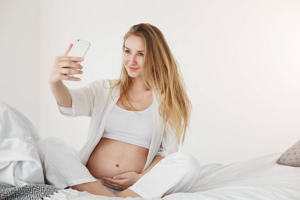 Prenatal selfie. Pregnant future mom making a self portrait using a smart phone smiling holding her belly - Photo, image