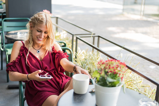 Щаслива блондинка красива жінка, використовуючи мобільний телефон, п'є чашку кави на терасі на відкритому повітрі
 - Фото, зображення