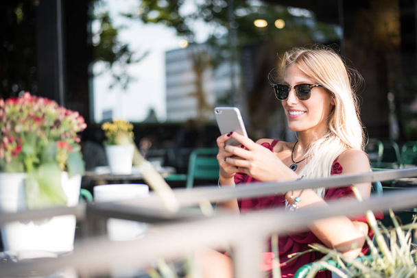 Feliz rubia hermosa mujer usando un teléfono móvil bebiendo una taza de café en una terraza al aire libre
 - Foto, Imagen