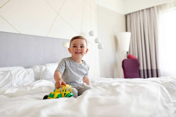 自宅やホテルのベッドでおもちゃの車を持った幸せな少年 - 写真・画像