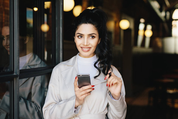 Horizontal portrait of brunette adorable woman in white clothes using her cell phone while standing outside having good mood touching her black curl of pony tail trying to make decision. Elegant girl - Фото, зображення