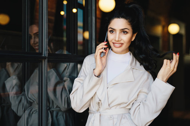 People, lifestyle, modern technologies and fashion concept. Portrait of fashionable woman with dark hair, bright eyes with long eyelashes and full lips wearing white jacket using smartphone outdoors - Φωτογραφία, εικόνα