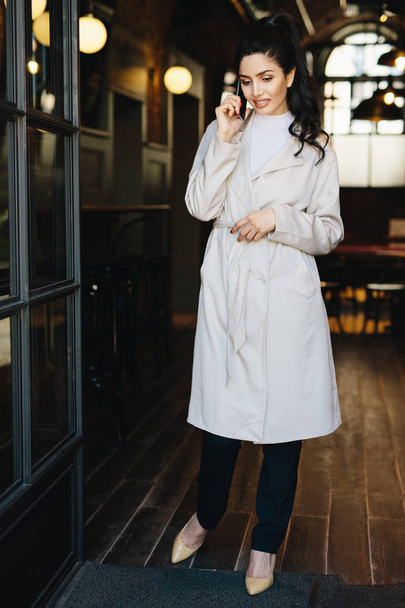 Ganzkörperporträt einer eleganten Geschäftsfrau in weißem Mantel, eleganten Schuhen und schwarzen Hosen, die im Café steht und ihren Geschäftspartner anruft, während er auf ihn wartet. Menschen, Technologiekonzept - Foto, Bild