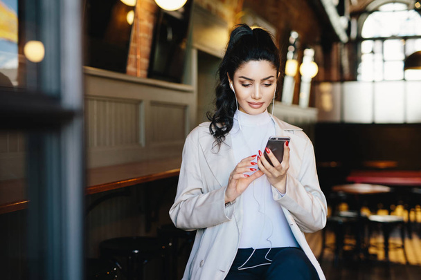 Kaunis brunette nainen poni hännän ja houkutteleva ulkonäkö istuu kahvilassa käyttämällä hänen älypuhelin surffailu internetissä ilmaiseksi wifi conncection ja kuunnella hänen suosikki musiikkia kuulokkeet
 - Valokuva, kuva