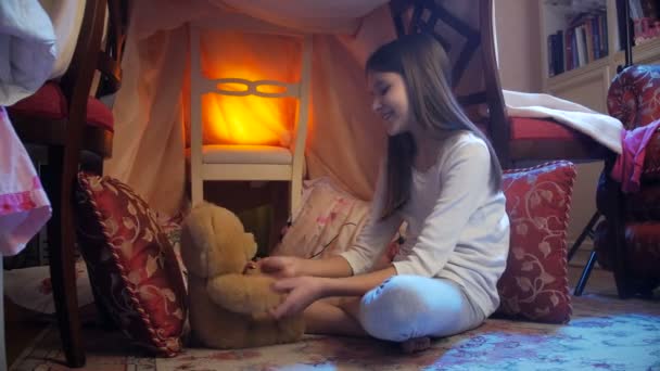 Dolly shot of cute smiling girl in pajamas playing with teddy bear in house made of blankets - Footage, Video