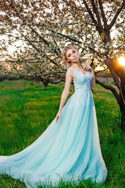 A girl in a long turquoise dress posing in the garden of blooming trees - 写真・画像
