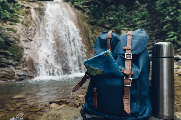 Hipster Blue Backpack, Map And Thermos Closeup. View From Front Tourist Traveler Bag On Waterfall Background. Adventure Hiking Concept - Foto, immagini