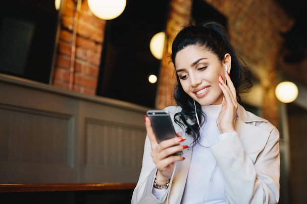 Χαμογελαστή γυναίκα με σκούρα μαλλιά δεμένα αλογοουρά φορώντας λευκά ρούχα κρατώντας smartphone ενώ ακούτε μουσική στο της ηλεκτρονικής συσκευής. Κομψή γυναίκα αναπαύεται στο café χρησιμοποιώντας app έχοντας χαμόγελο στο πρόσωπό - Φωτογραφία, εικόνα
