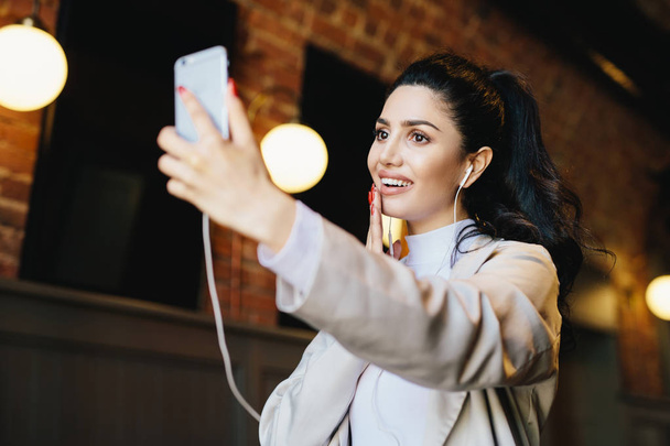 Brunette woman in white jacket having dark eyes with long eyelashes, full lips and healthy skin having good mood while resting in cafe listening to different tracks with earphones and making selfie - Photo, Image