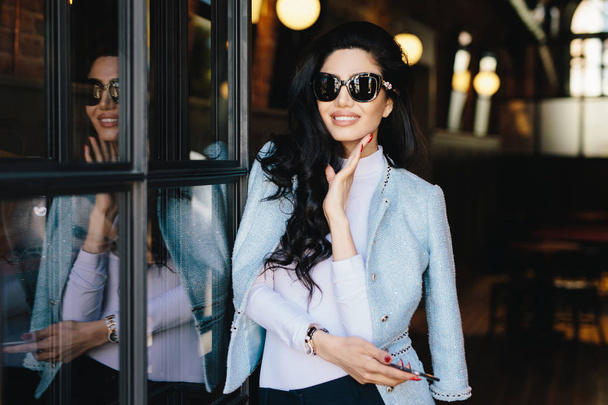 Retrato de una joven glamurosa con cabello ondulado oscuro con gafas de sol y blusa blanca con chaqueta elegante con manicura roja en el interior sosteniendo el teléfono inteligente esperando a su novio en la cafetería
 - Foto, Imagen