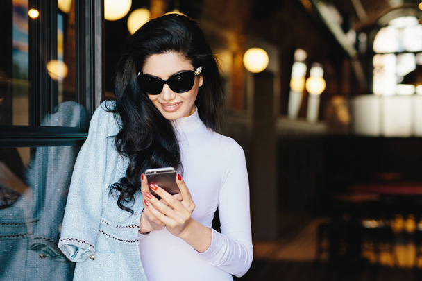 Muhteşem esmer kadın beyaz giysiler ve ücretsiz internet conncection kullanarak arkadaşlarınızla mesajlaşma onun smartphone kullanarak kafede oturan güneş gözlüğü yatay portresi. Güzellik ve gençlik kavramı - Fotoğraf, Görsel