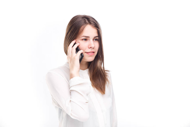 Beautiful young girl in a white shirt on white isolated background talking on a mobile phone. Smiles portrait to the waist - 写真・画像