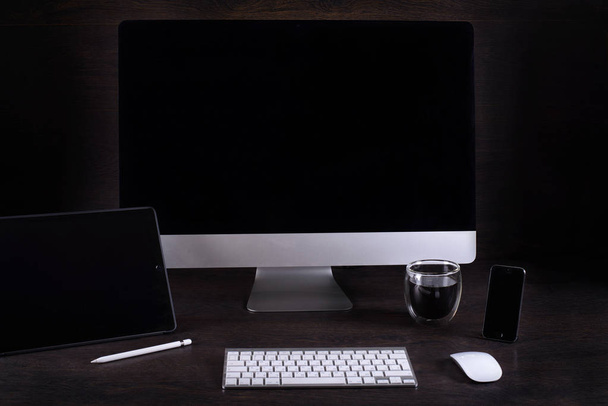 черный экран компьютера в комнате на коричневом столе и кинокамера, кофейная кружка, телефон, планшетный компьютер, напротив темной стены
 - Фото, изображение