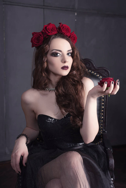 красива молода жінка з квіткою троянд у волоссі, одягнена в чорну сукню з макіяжем на темному тлі, готичною атмосферою. темно-червоні губи. крупним планом модний ретуш портрет
 - Фото, зображення