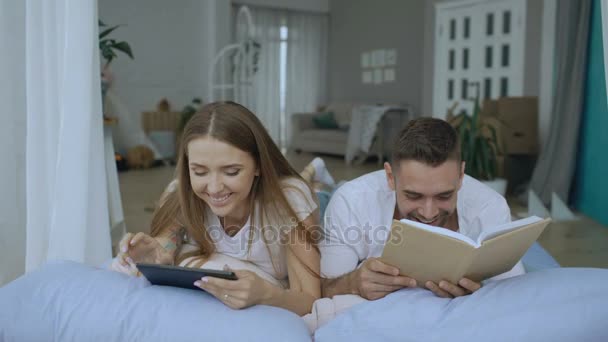 Young couple have leisure time lying in bed and reading book and tablet computer at home - 映像、動画
