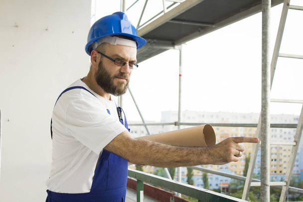 Chybné stavební dělník v pracovním oblečení a helmu stojí ve vysoké nadmořské výšce na staveništi s plány pod paží a vztekle ukazuje prstem. - Fotografie, Obrázek
