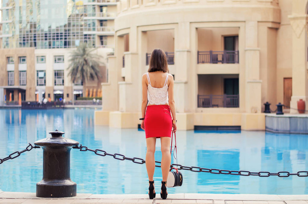 Mulher europeia magro e alto em top de cultura branca e saia vermelha curta com saco pequeno e sapatos de salto alto em pé perto da orla no centro de Dubai
 - Foto, Imagem