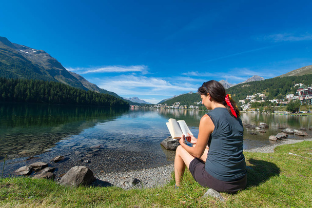 Nuori nainen lukee kirjaa vuoristojärven edessä.
 - Valokuva, kuva