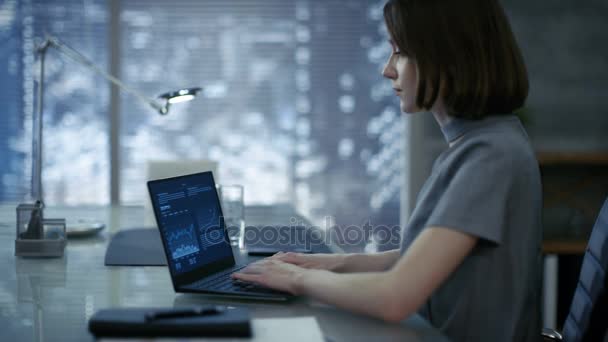Beautiful, Stylishly Dressed Young Businesswoman Sits at Her Working Table and Types on a Laptop. Her Office Looks Modern with Dark Overtones, Big City Behind the Window. - Footage, Video