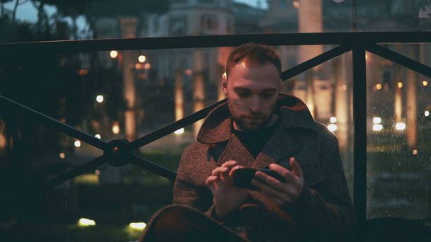 Young handsome man using smartphone, evening city on a background. Man browse the Internet with touchscreen technology. - Φωτογραφία, εικόνα