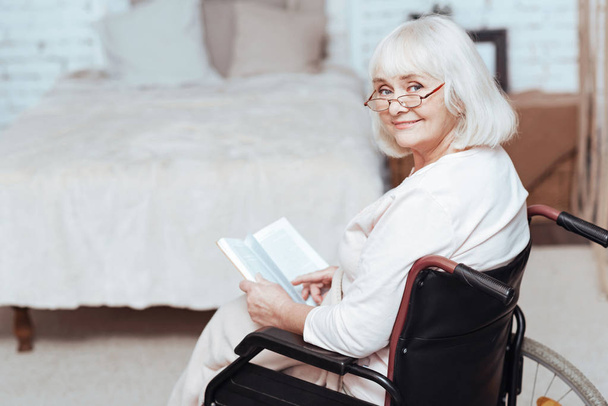 Positive retired woman reading book in wheelchair at home - Foto, immagini