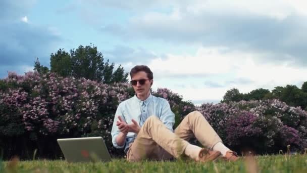 Молодий чоловік має відеодзвінок на ноутбуці, сидячи на зеленій траві в парку. Літній день
 - Кадри, відео