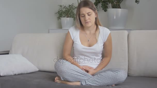 Smutny młoda kobieta cierpi na okres boleć siedzi na kanapie i trzymając się za ręce na brzuchu - Materiał filmowy, wideo