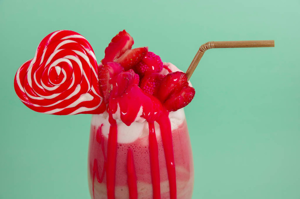 Delicious homemade extreme milkshake of strawberry with milk foam with a plastic straw and a pink blackberry candy with a heart candy on top, in a soft blue background - Zdjęcie, obraz