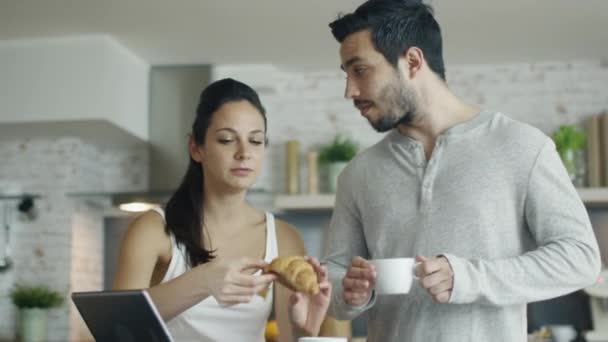 Handsome Couple in the Kitchen with Tablet on the Table. She eats Croissant He Drinks Coffee. - Video