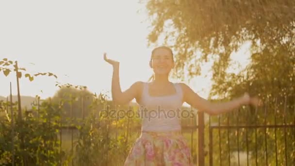 Ευτυχισμένος νεαρή γυναίκα που χορεύουν κάτω από το νερό από κήπο ψεκαστήρων στο ηλιοβασίλεμα - Πλάνα, βίντεο