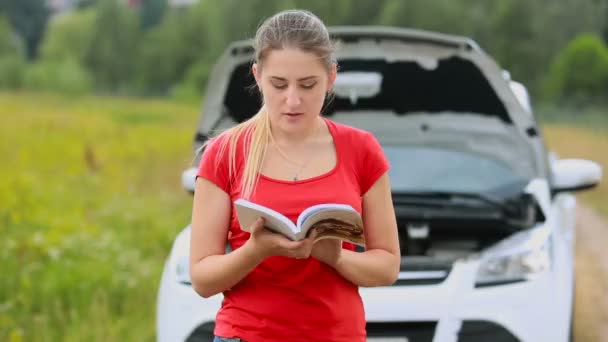 Joven mujer confundida de pie en el coche brokedown en el campo y leer el manual del usuario
 - Metraje, vídeo