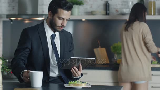 Handsome Businessman Uses Tablet Computer while Having Breakfast at His Kitchen while His Girlfriend Cooks in the Background. - Materiaali, video
